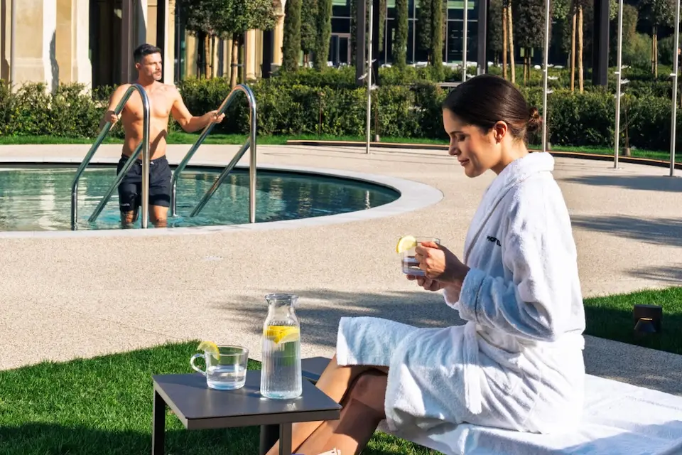 Coppia che si gode un giorno alla spa; la donna si rilassa in accappatoio con un drink, l'uomo nuota in piscina. Benessere.