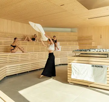 Interno sauna in legno chiaro con donne rilassate e persona che sventola asciugamano. DE-MONTEL