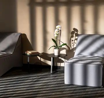 Interno con poltrone grigie, tavolino con orchidea e libri, illuminato da luce filtrata.