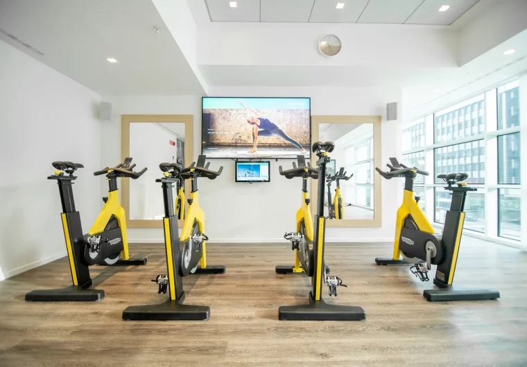 Cyclette da spinning gialle e nere in una sala fitness luminosa con schermo.